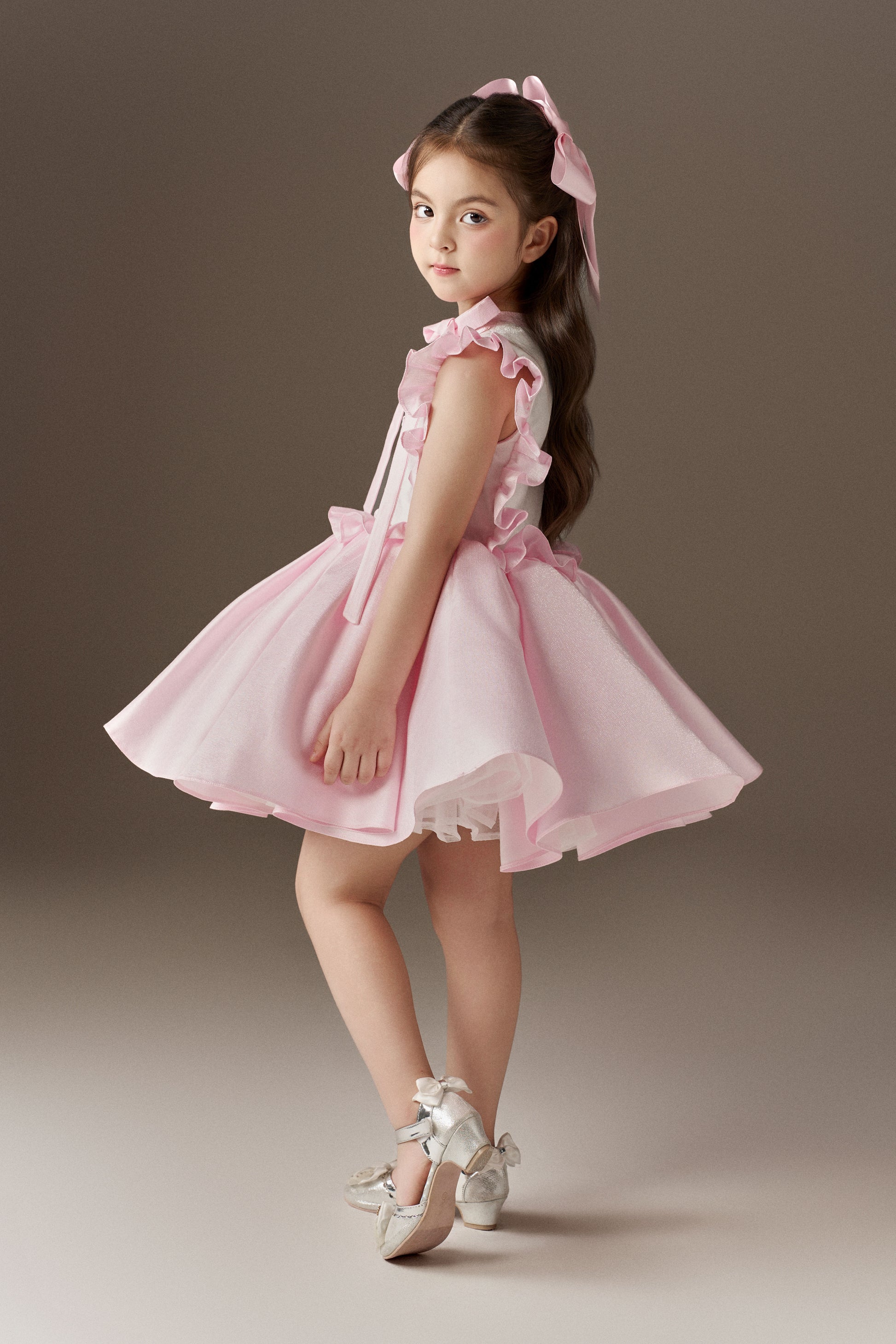 A side view of a pink sleeveless children's dress, showcasing its voluminous skirt and large ruffle details. The bodice is decorated with hand-applied beads and sequins, and two oversized pink hair bows are worn.
