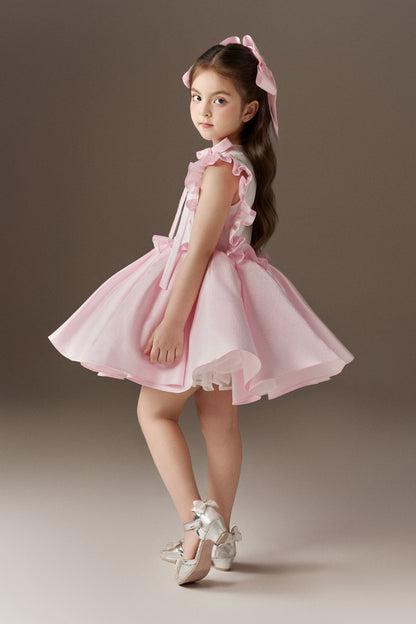 A side view of a pink sleeveless children's dress, showcasing its voluminous skirt and large ruffle details. The bodice is decorated with hand-applied beads and sequins, and two oversized pink hair bows are worn.