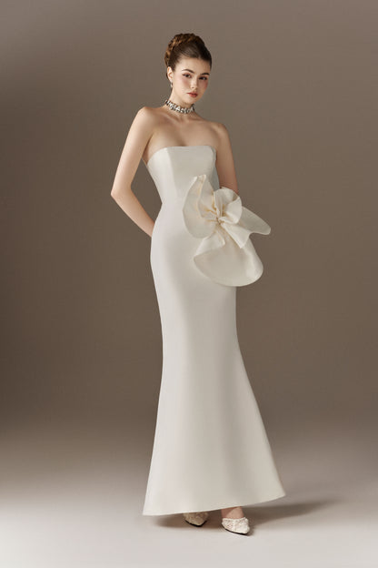 A model poses in a white strapless fit-and-flare maxi dress featuring an oversized 3D flower detail on the left hip, looking slightly to her right.