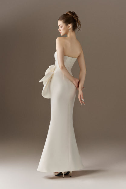 The back view of a strapless white mermaid-style maxi dress, showcasing the silhouette and a large, structured floral embellishment visible on the left hip from the rear.