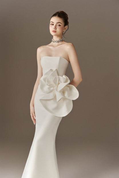  A strapless white mermaid-style maxi dress featuring a prominent, oversized three-dimensional floral embellishment on the left hip.