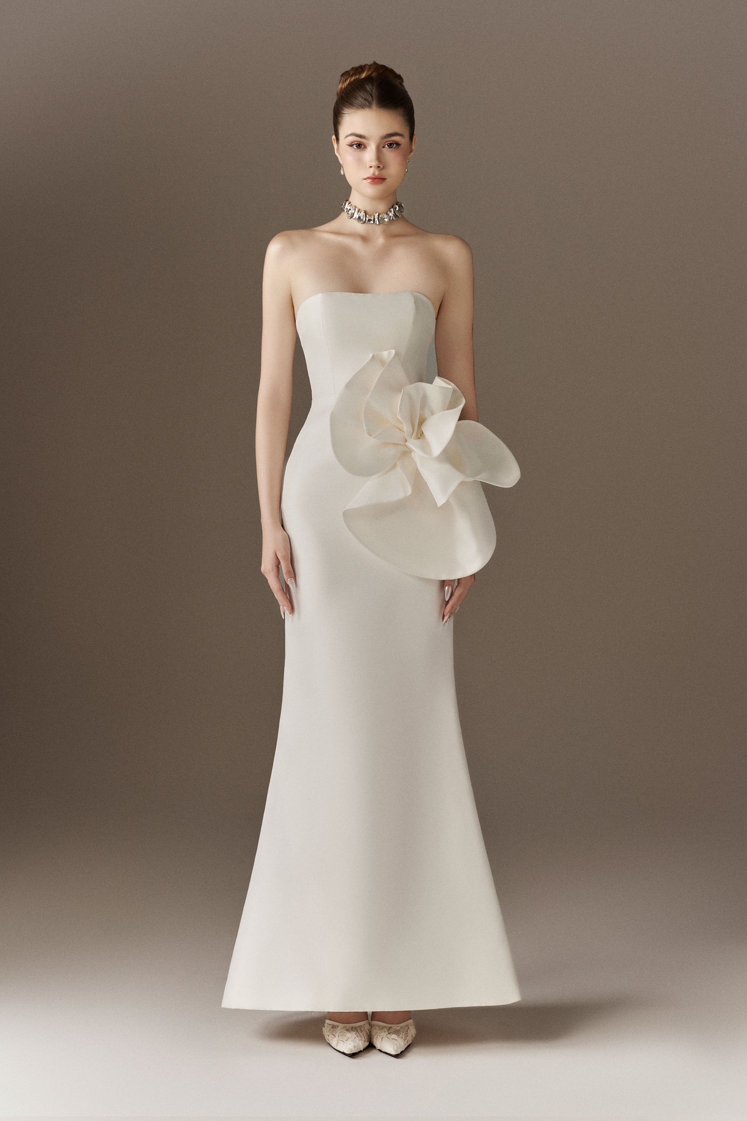 A white strapless maxi dress, highlighting its bandeau neckline, figure-hugging fit, and the oversized 3D handcrafted floral embellishment on the left hip. The dress flares out at the bottom.