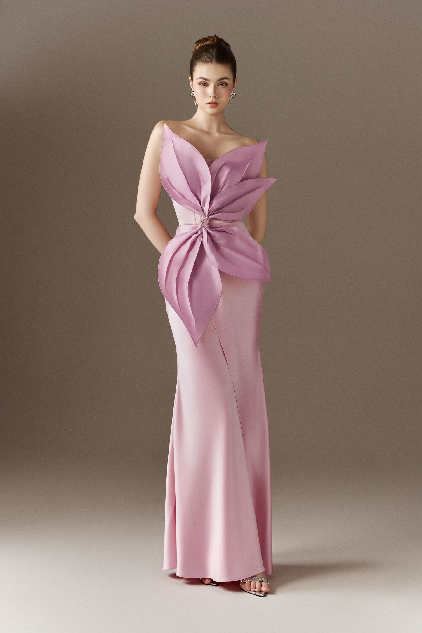  A pink, sleeveless, figure-hugging maxi dress with a bandeau neckline and an off-shoulder open neckline. The dress features a large 3D handcrafted petal detail at the waist and a front slit.
