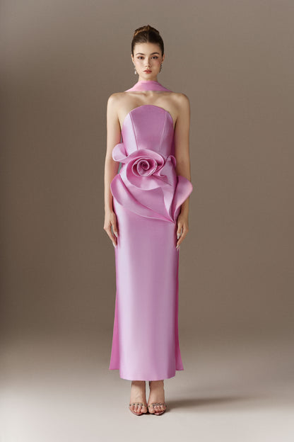 A vibrant pink strapless maxi dress featuring a bandeau neckline and a large, handcrafted three-dimensional rose embellishment draped on the left hip.
