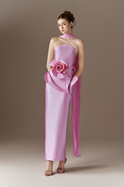 A close-up view of a pink strapless maxi dress, highlighting the bandeau neckline, the fitted bodice, and an elaborate, oversized 3D rose embellishment on the left hip, with a long, narrow scarf draped over the right shoulder.