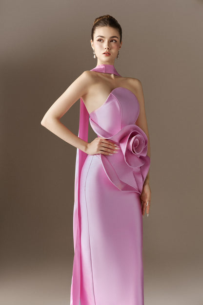 A pink strapless maxi dress with a fitted bodice, a large draped 3D rose accent on the hip, and a long, matching detachable cape extending from the left side.