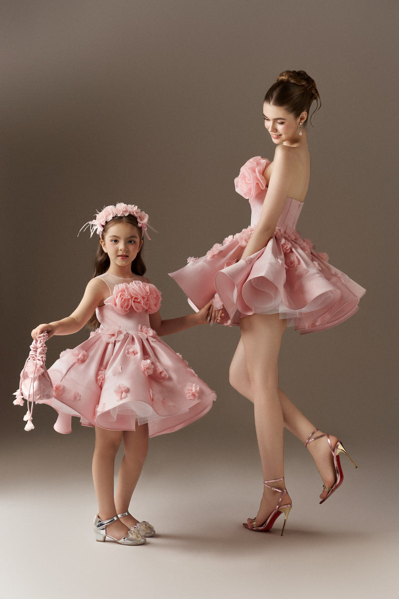 Two matching pink dresses, one adult-sized and one child-sized. The adult dress is strapless with a ruffled bodice and a voluminous sculpted skirt with floral embellishments. The child's dress has spaghetti straps, a sheer mesh neckline, a ruffled bodice, and a voluminous sculpted skirt with floral embellishments. A child's handbag and floral headband are included.